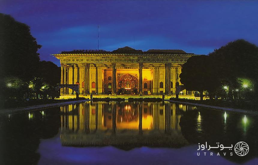کاخ چهلستون اصفهان در شب که با نوری زردرنگ روشن شده و حوضی جلوی آن است. 